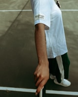 A person is standing on a court, wearing a white shirt with the words 'khoman room' on the sleeve. They are wearing dark pants and holding onto a tennis net. Only one arm is visible, and the background is a blurred tennis court surface.