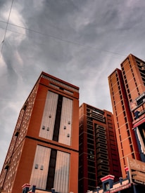 Tall modern buildings with a reddish-brown color dominate the scene against a cloudy sky. The structures have large glass windows and clean lines, giving a sense of urban development and architectural design. There are power lines visible in front of the buildings.