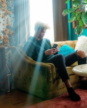 A person with curly hair is sitting on a vintage-style sofa in a cozy, retro-themed room. They are looking at a smartphone. Soft natural light streams in through a window, highlighting floral wallpaper and houseplants.