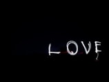 The word 'LOVE' is written in light against a dark background, likely using a long-exposure photography technique. The letters are illuminated and stand out in contrast to the black space around them.