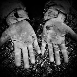 Two open hands are shown covered in dirt and grime. The hands are positioned palm-facing up and appear rough and worn, indicating manual labor or outdoor work. The background is a textured surface, possibly concrete or pavement.