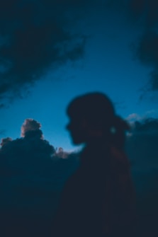 A silhouette of a person appears against a backdrop of dark, dramatic clouds with a blue sky. The image is atmospheric, creating a sense of mystery and introspection.