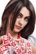 A woman with dark hair is adorned with a traditional embroidered garment featuring red floral patterns. She is holding the fabric delicately and wearing a string of red beads around her neck.