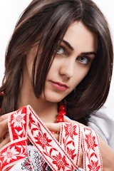 A woman with dark hair is adorned with a traditional embroidered garment featuring red floral patterns. She is holding the fabric delicately and wearing a string of red beads around her neck.