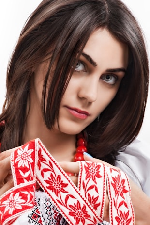 A woman with dark hair is adorned with a traditional embroidered garment featuring red floral patterns. She is holding the fabric delicately and wearing a string of red beads around her neck.