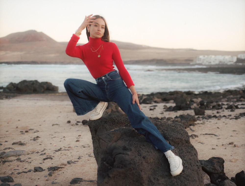 woman in red long sleeve shirt and blue denim jeans sitting on rock during daytime