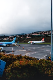 Two commercial airplanes are parked on an airport tarmac. In the foreground, there are some airport workers wearing high-visibility vests walking near the planes. In the background, there is a hillside with houses nestled among greenery, under a cloudy sky.