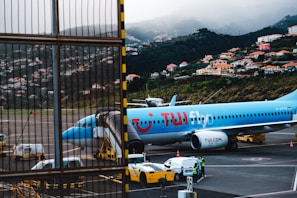 An airplane from TUI is parked on an airport tarmac, positioned near a jet bridge for boarding. Ground support vehicles and personnel are visible nearby. The backdrop features a scenic hillside dotted with numerous houses and buildings, with misty clouds partially obscuring the hilly landscape.