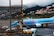 An airplane from TUI is parked on an airport tarmac, positioned near a jet bridge for boarding. Ground support vehicles and personnel are visible nearby. The backdrop features a scenic hillside dotted with numerous houses and buildings, with misty clouds partially obscuring the hilly landscape.