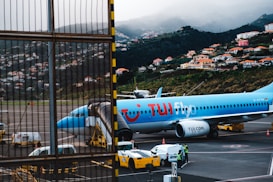 An airplane from TUI is parked on an airport tarmac, positioned near a jet bridge for boarding. Ground support vehicles and personnel are visible nearby. The backdrop features a scenic hillside dotted with numerous houses and buildings, with misty clouds partially obscuring the hilly landscape.