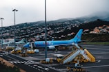 An airport scene featuring a blue TUI Fly passenger plane on the tarmac. The aircraft is connected to a boarding stairway with several ground support vehicles and personnel nearby. In the background, a hillside is covered with numerous small houses, partially obscured by mist or fog.
