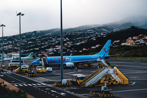 An airport scene featuring a blue TUI Fly passenger plane on the tarmac. The aircraft is connected to a boarding stairway with several ground support vehicles and personnel nearby. In the background, a hillside is covered with numerous small houses, partially obscured by mist or fog.
