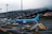 An airport scene featuring a blue TUI Fly passenger plane on the tarmac. The aircraft is connected to a boarding stairway with several ground support vehicles and personnel nearby. In the background, a hillside is covered with numerous small houses, partially obscured by mist or fog.