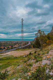 A scenic landscape features a tall transmission tower in the center, surrounded by a lush, green hillside dotted with rocks and plants. A road runs through the middle ground, leading towards a distant horizon where the land meets the sea under a cloudy, overcast sky.