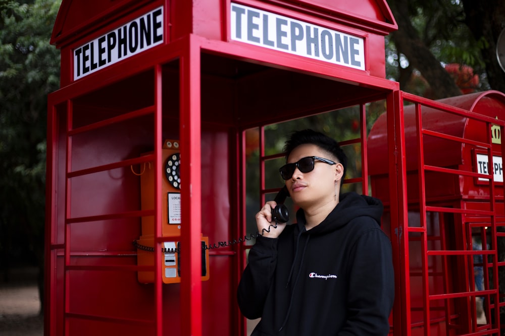 homem de óculos escuros pretos e capuz preto de pé perto da cabine telefônica vermelha