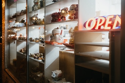 A display window filled with various ceramic and porcelain figurines is illuminated by artificial lighting. Among these items, several large, decorative cat figurines are prominent. A neon 'OPEN' sign glows brightly on the right-hand side, contributing to a warm and inviting atmosphere.