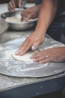 Hands are seen pressing and shaping dough on a floured surface, possibly part of a baking or pizza preparation process. A metal bowl in the background contains more dough, indicating ongoing food preparation.