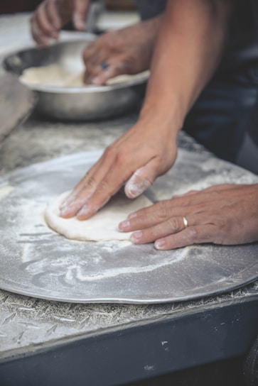 Hands are seen pressing and shaping dough on a floured surface, possibly part of a baking or pizza preparation process. A metal bowl in the background contains more dough, indicating ongoing food preparation.