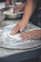 Hands are seen pressing and shaping dough on a floured surface, possibly part of a baking or pizza preparation process. A metal bowl in the background contains more dough, indicating ongoing food preparation.