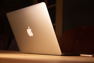 A sleek silver laptop with a glowing logo on its lid, placed on a wooden surface. The lighting is warm and subdued, creating a cozy ambiance.