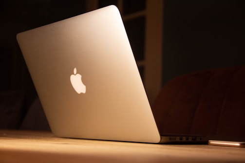 A sleek silver laptop with a glowing logo on its lid, placed on a wooden surface. The lighting is warm and subdued, creating a cozy ambiance.