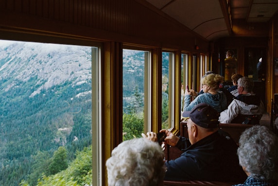 A group of people are sitting inside a train carriage, looking out the large windows at a scenic view of a mountainous landscape. Some individuals are taking photos or gazing at the view, indicating a sightseeing experience. The interior of the train has wooden paneling and a cozy atmosphere.
