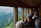 A group of people are sitting inside a train carriage, looking out the large windows at a scenic view of a mountainous landscape. Some individuals are taking photos or gazing at the view, indicating a sightseeing experience. The interior of the train has wooden paneling and a cozy atmosphere.