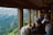 A group of people are sitting inside a train carriage, looking out the large windows at a scenic view of a mountainous landscape. Some individuals are taking photos or gazing at the view, indicating a sightseeing experience. The interior of the train has wooden paneling and a cozy atmosphere.