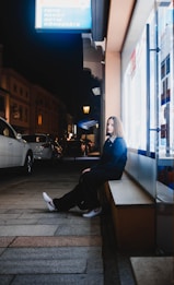 A young person sits on a bench outside a well-lit storefront at night. The street is lined with parked cars and glowing streetlamps that add a serene, urban setting. The person is casually dressed, and the illuminated sign above suggests the shop offers various services or products.