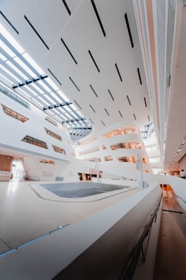 A modern architectural interior with sleek, angular designs and large skylights allowing natural light to flood the space. The walls and ceiling are predominantly white, with some brown and gray accents. The flooring appears to be a polished, light-colored surface, and there are multiple levels with walkways and railings.