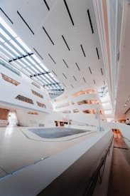 A modern architectural interior with sleek, angular designs and large skylights allowing natural light to flood the space. The walls and ceiling are predominantly white, with some brown and gray accents. The flooring appears to be a polished, light-colored surface, and there are multiple levels with walkways and railings.