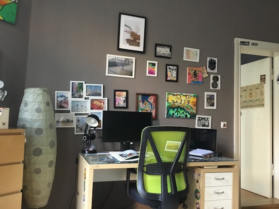 A cozy workspace featuring a desk with a computer monitor, a green office chair, and various papers on the desk. The wall behind is decorated with numerous small framed pictures and art pieces, giving a creative and personalized look. To the left, there is a tall lamp with a polka-dot pattern and a set of drawers partially visible.