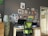 A cozy workspace featuring a desk with a computer monitor, a green office chair, and various papers on the desk. The wall behind is decorated with numerous small framed pictures and art pieces, giving a creative and personalized look. To the left, there is a tall lamp with a polka-dot pattern and a set of drawers partially visible.
