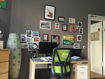 A cozy workspace featuring a desk with a computer monitor, a green office chair, and various papers on the desk. The wall behind is decorated with numerous small framed pictures and art pieces, giving a creative and personalized look. To the left, there is a tall lamp with a polka-dot pattern and a set of drawers partially visible.