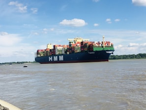 A large cargo ship with the letters HMM on its side is sailing on a wide river. The vessel is loaded with numerous colorful shipping containers. The sky is bright with scattered white clouds, and there's a line of trees in the background along the riverbank.