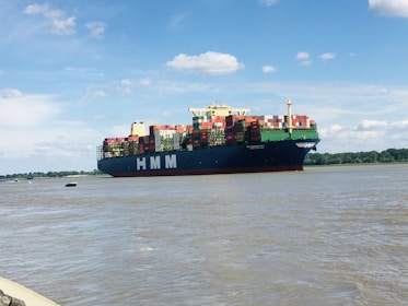 A large cargo ship with the letters HMM on its side is sailing on a wide river. The vessel is loaded with numerous colorful shipping containers. The sky is bright with scattered white clouds, and there's a line of trees in the background along the riverbank.