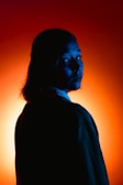 A person with long hair is silhouetted against a vibrant orange and red background. The use of blue lighting on the face creates a stark contrast, bringing out the facial features in a dramatic manner.