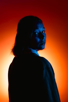 A person with long hair is silhouetted against a vibrant orange and red background. The use of blue lighting on the face creates a stark contrast, bringing out the facial features in a dramatic manner.