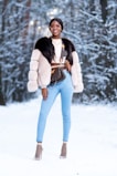 A person stands confidently in a snowy forest wearing stylish winter attire, including a fur coat and light blue jeans. They accessorize with sunglasses, layered necklaces, and high heels with netted socks. The background features snow-covered trees creating a winter wonderland setting.