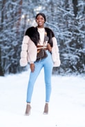 A person stands confidently in a snowy forest wearing stylish winter attire, including a fur coat and light blue jeans. They accessorize with sunglasses, layered necklaces, and high heels with netted socks. The background features snow-covered trees creating a winter wonderland setting.