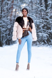 A person stands confidently in a snowy forest wearing stylish winter attire, including a fur coat and light blue jeans. They accessorize with sunglasses, layered necklaces, and high heels with netted socks. The background features snow-covered trees creating a winter wonderland setting.