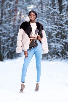 A person stands confidently in a snowy forest wearing stylish winter attire, including a fur coat and light blue jeans. They accessorize with sunglasses, layered necklaces, and high heels with netted socks. The background features snow-covered trees creating a winter wonderland setting.