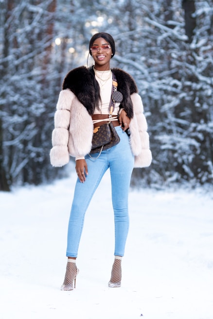 A person stands confidently in a snowy forest wearing stylish winter attire, including a fur coat and light blue jeans. They accessorize with sunglasses, layered necklaces, and high heels with netted socks. The background features snow-covered trees creating a winter wonderland setting.
