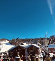 A bustling ski resort scene features a wooden building adorned with sculptures and surrounded by snow-covered mountains. People are gathered outside, enjoying the sunny day, while ski slopes and a clear blue sky are visible in the background.
