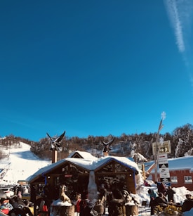 A bustling ski resort scene features a wooden building adorned with sculptures and surrounded by snow-covered mountains. People are gathered outside, enjoying the sunny day, while ski slopes and a clear blue sky are visible in the background.