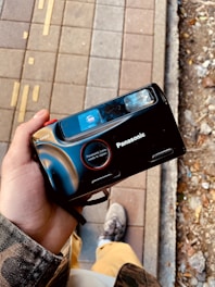 A person holding a black Panasonic camera with a vintage design. The camera features a prominent lens labeled as 'Panasonic Lens made in Japan'. The background shows a pavement with tactile indicators and nearby soil with scattered debris.