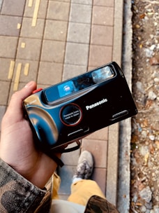 A person holding a black Panasonic camera with a vintage design. The camera features a prominent lens labeled as 'Panasonic Lens made in Japan'. The background shows a pavement with tactile indicators and nearby soil with scattered debris.
