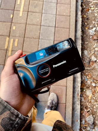 A person holding a black Panasonic camera with a vintage design. The camera features a prominent lens labeled as 'Panasonic Lens made in Japan'. The background shows a pavement with tactile indicators and nearby soil with scattered debris.