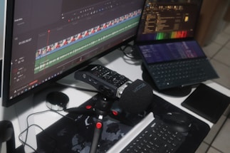 A workspace setup featuring a large monitor displaying video editing software with audio and video tracks visible on the timeline. A digital audio recorder with a foam windscreen is mounted on a small tripod in front of a keyboard. A laptop displaying various software interfaces is positioned nearby, along with a computer mouse and a tablet.