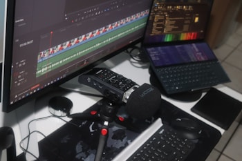 A workspace setup featuring a large monitor displaying video editing software with audio and video tracks visible on the timeline. A digital audio recorder with a foam windscreen is mounted on a small tripod in front of a keyboard. A laptop displaying various software interfaces is positioned nearby, along with a computer mouse and a tablet.